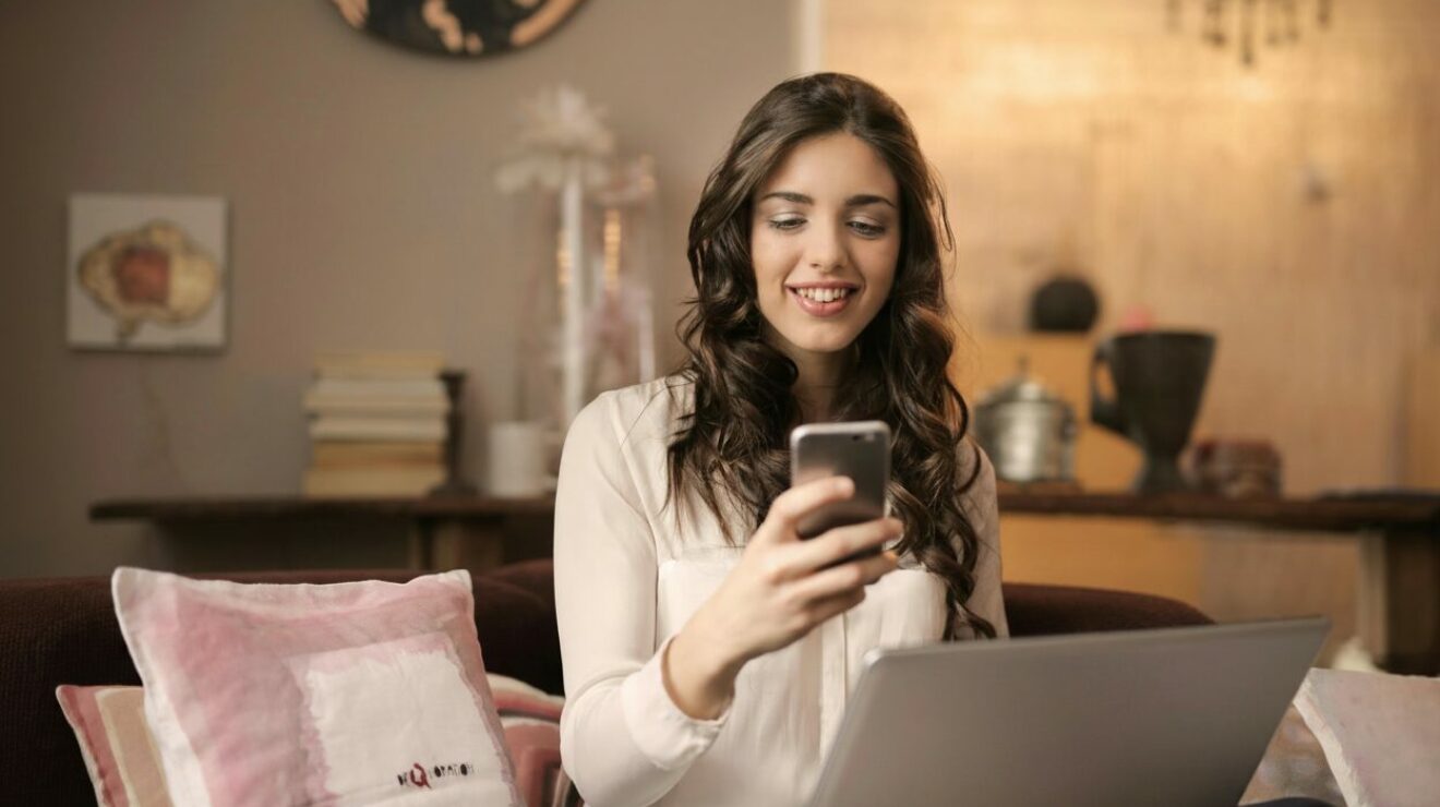 Frau mit Smartphone vor Notebook in Wohnzimmerumgebung.