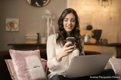 Frau mit Smartphone vor Notebook in Wohnzimmerumgebung.